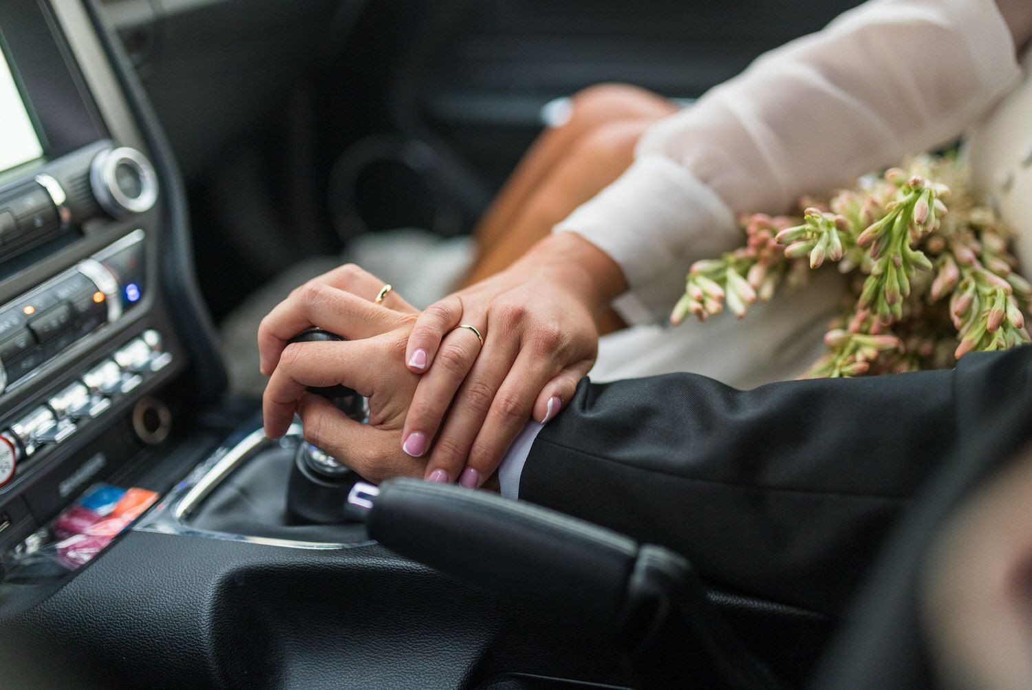 Pareja tomada de la mano en un coche, la mano de la mujer encima con el anillo de bodas, flores cerca.