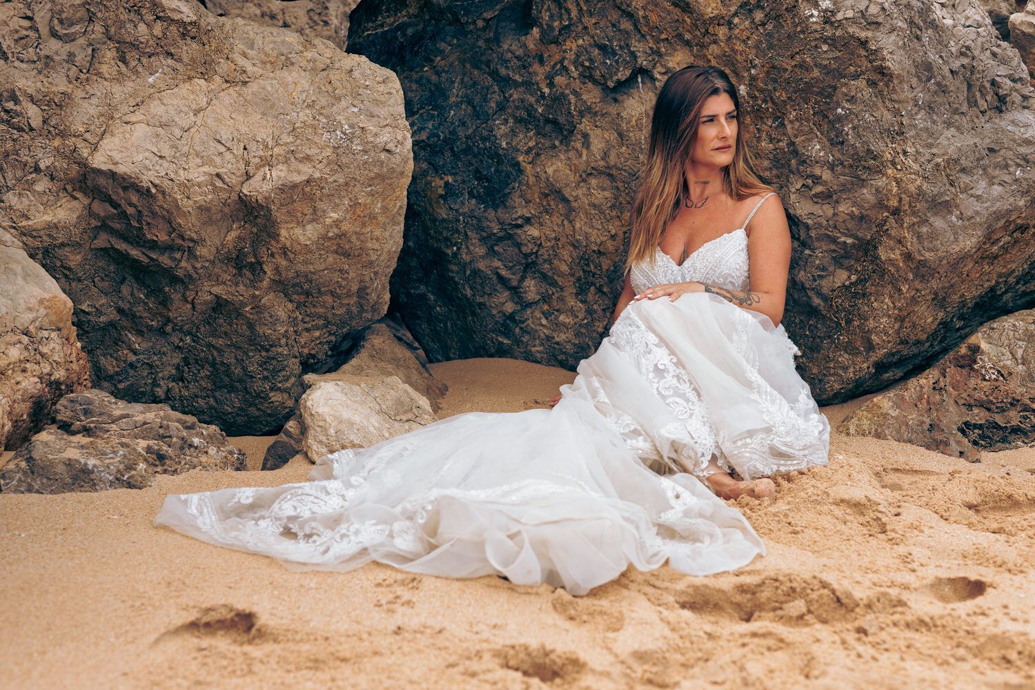 Una mujer con un vestido de novia blanco se sienta en la playa, mirando hacia otro lado, cerca de las rocas.
