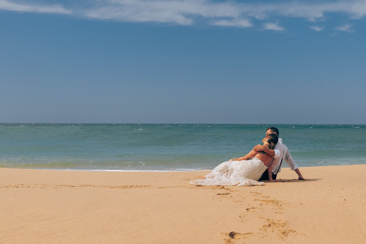 Los novios se sientan en una playa de arena, con el océano y el cielo azul de fondo.