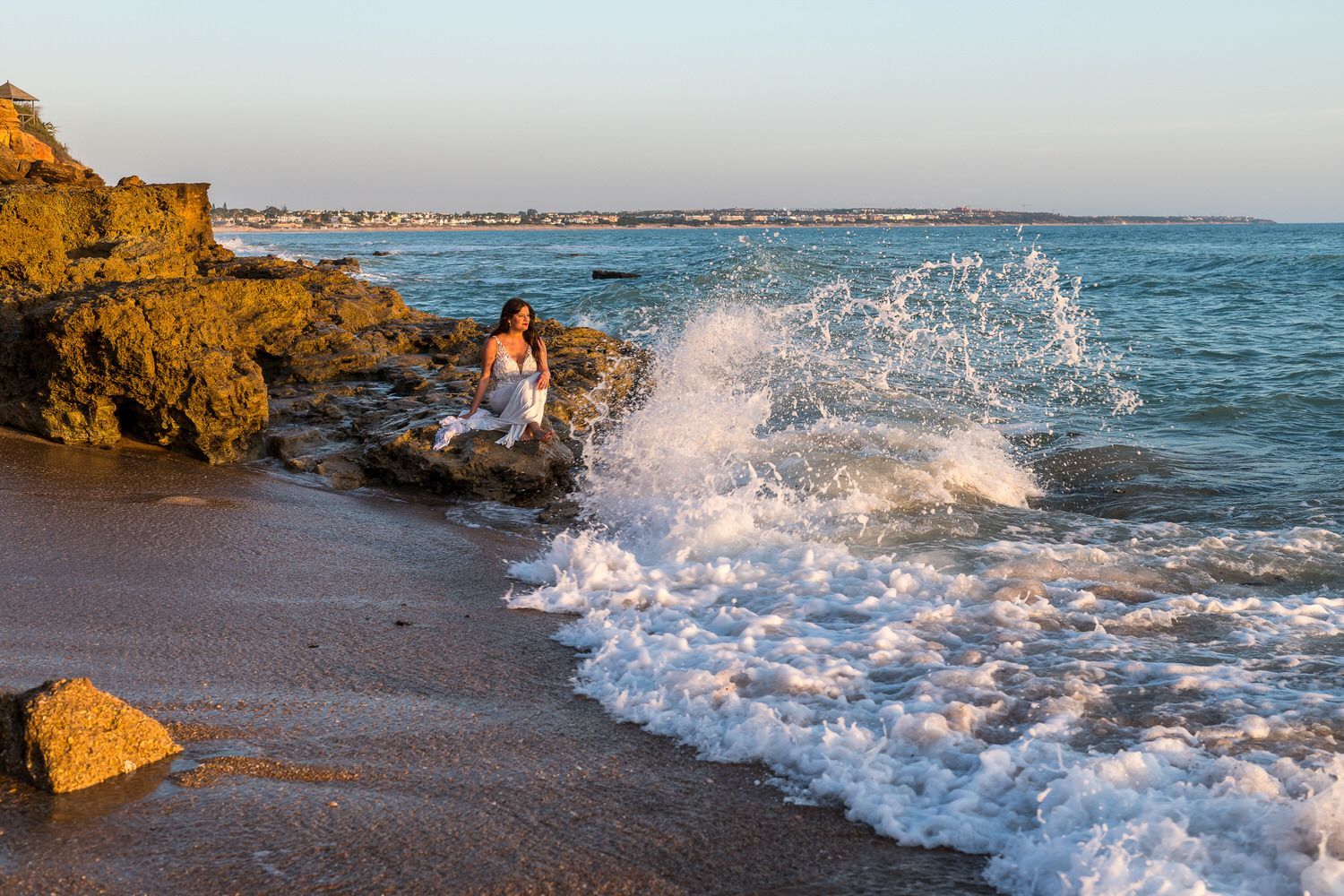 Una mujer vestida de blanco se sienta en las rocas mientras las olas del océano salpican, con la playa al fondo.