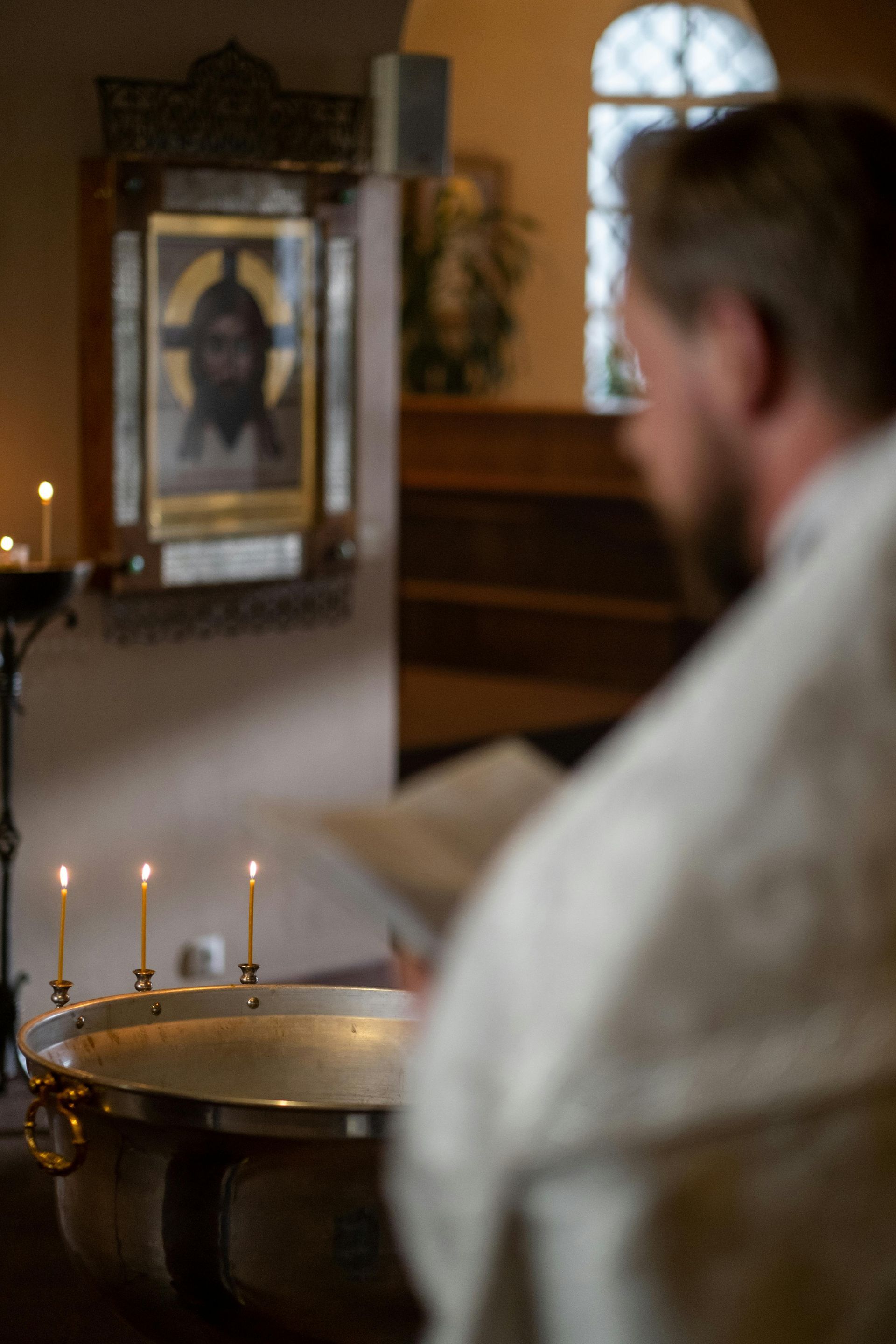Sacerdote con túnica blanca cerca de una pila bautismal con velas encendidas, icono al fondo.