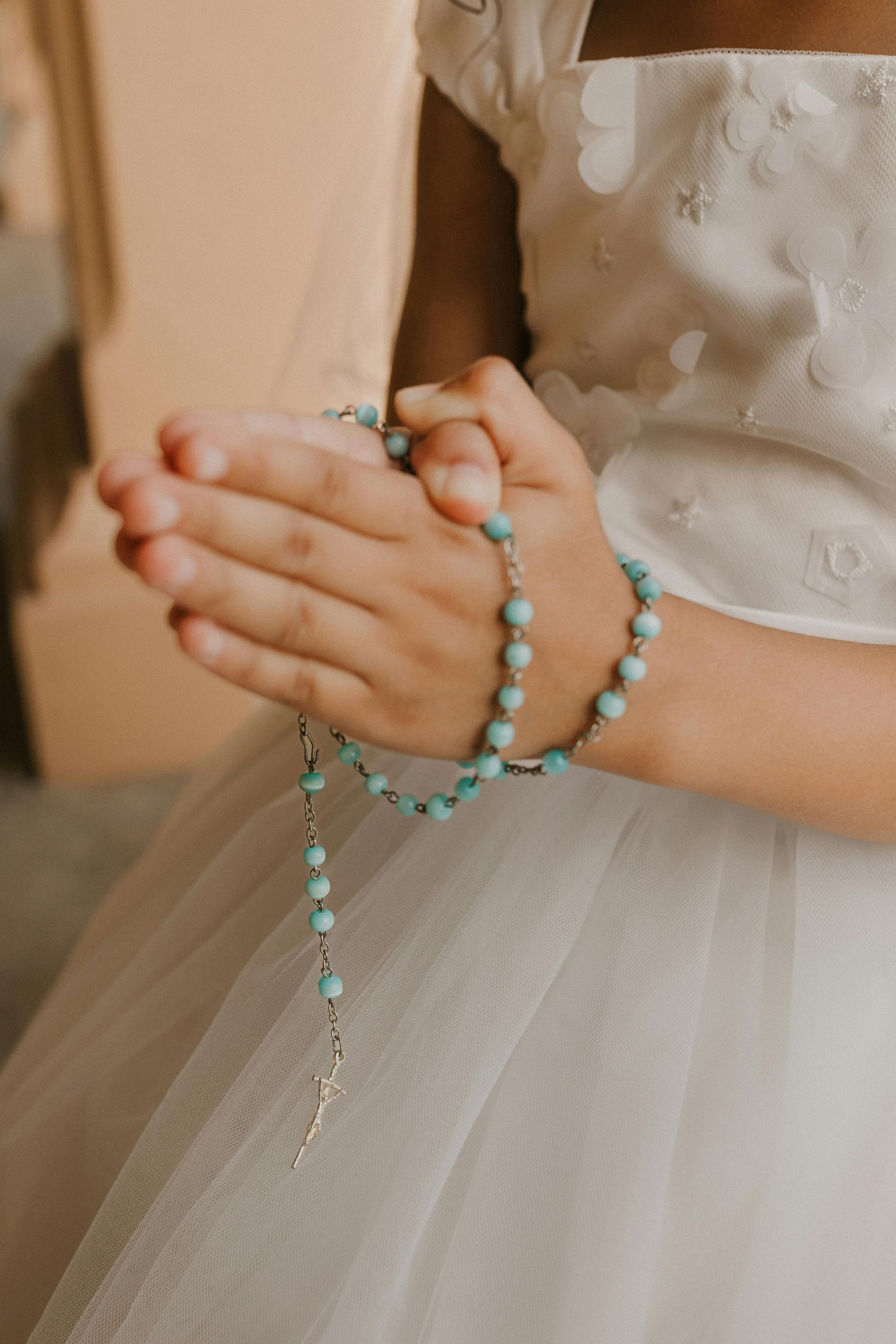 Niño con vestido blanco sosteniendo un rosario turquesa, con las manos juntas en oración.