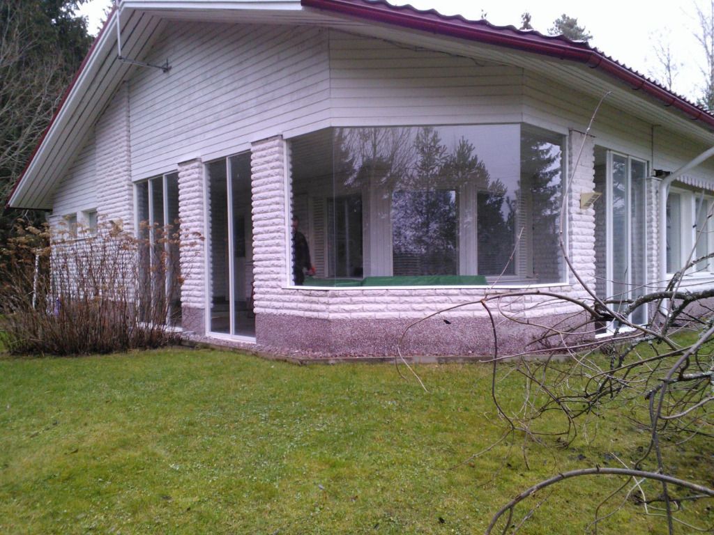 Valkoinen talo, jossa on suuret ikkunat, joista on näkymä pihalle, jossa on vihreää ruohoa ja puita.