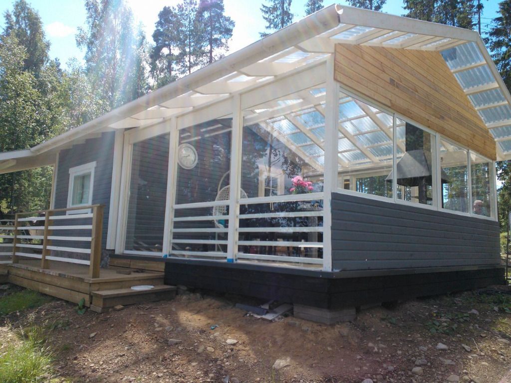 Mökki lasiseinäisellä kuistilla; harmaa ulkoverhous, valkoinen lista, puinen kansi. Metsää taustalla.