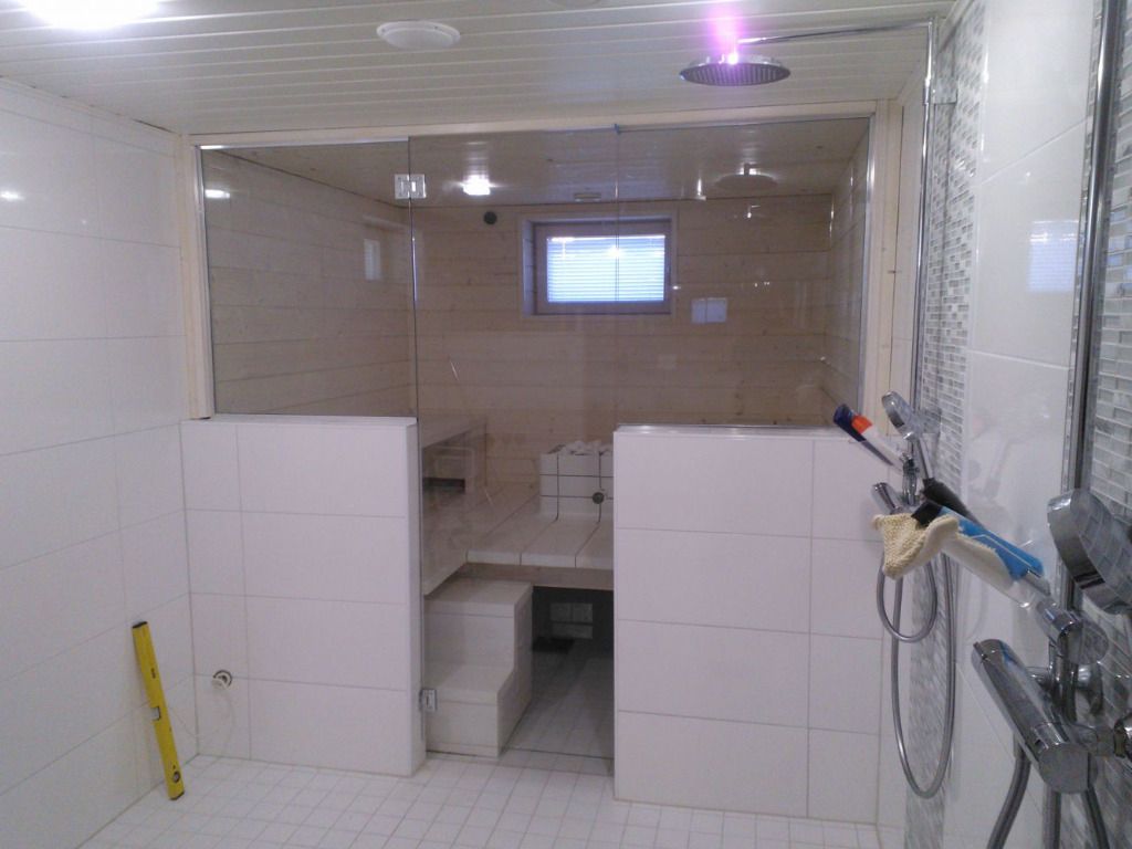 Valkoiseksi laatoitettu höyrysaunan sisustus, jossa on lasiseinät ja ikkuna, johon pääsee oviaukon kautta.