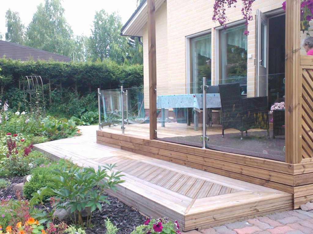 Puinen terassi lasikaiteella, jolta on näkymä puutarhaan ja talolle. Kukkia ja vehreyttä ympäröi.