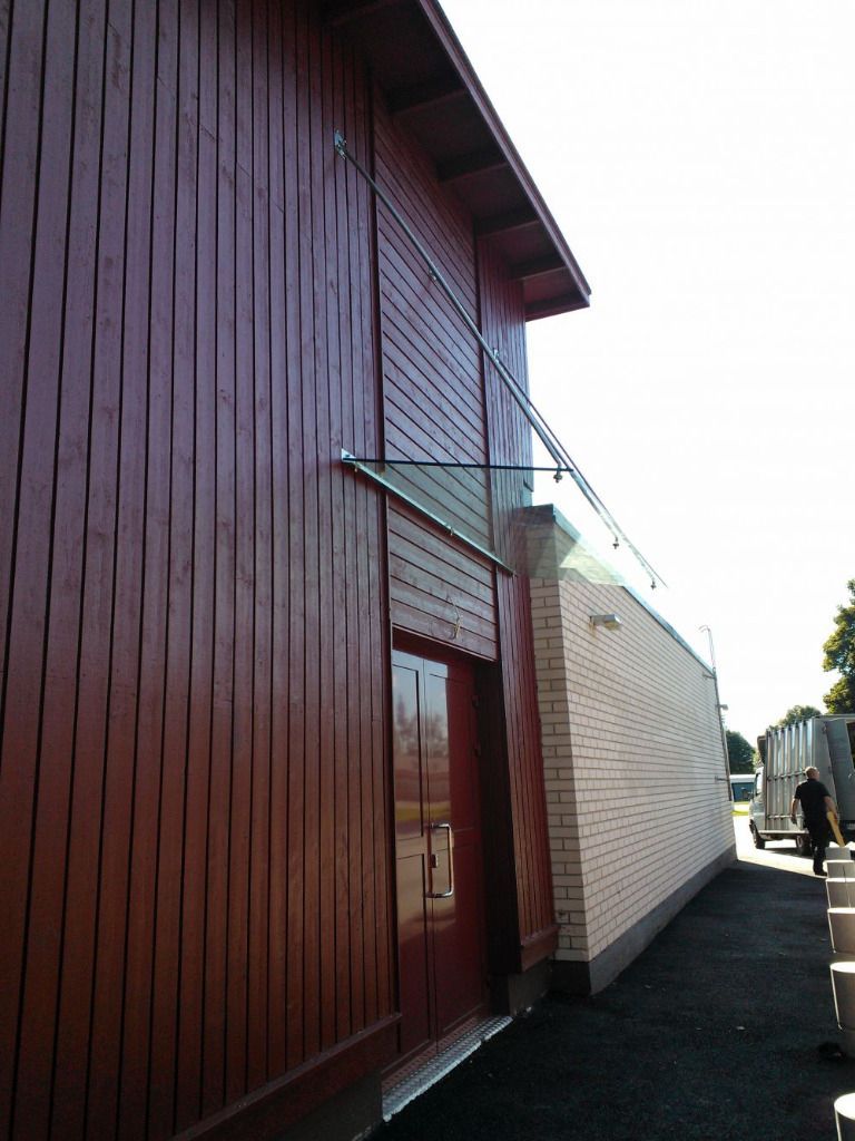 Punainen puinen rakennus, jossa on lasikatos, valkoisen tiiliseinän vieressä. Henkilö pakettiauton lähellä oikealla.