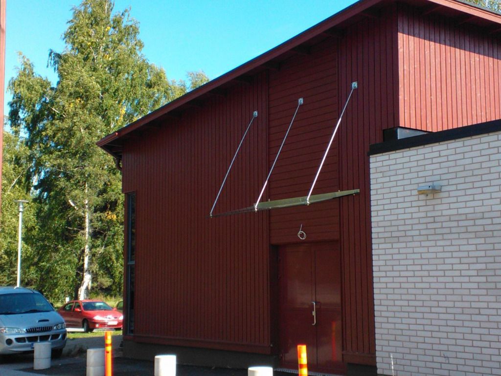 Punainen puurakennus, jossa on katos, ovi ja valkoinen tiiliseinä oikealla. Autoja pysäköitynä vasemmalle.