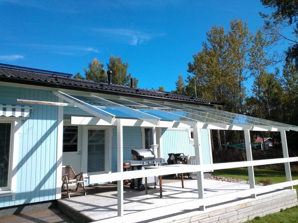 Sininen talo, jossa valkoinen kuisti ja läpinäkyvä katto, grilli ja ulkoterassi. Aurinkoinen päivä.