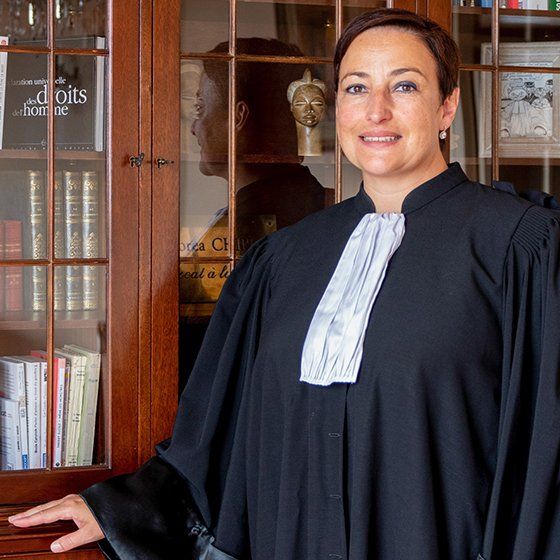 Maître Lorea CHIPI en robe d'avocat, souriante, se tenant debout devant sa bibliothèque