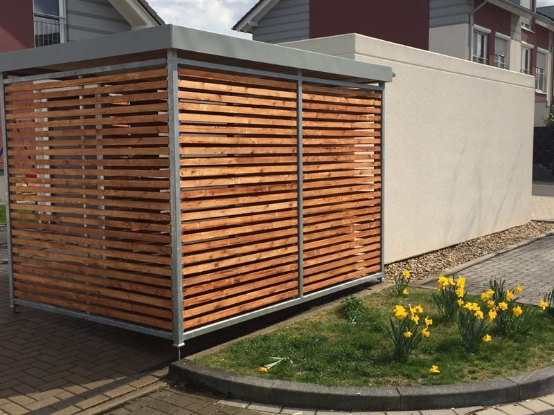 Ein Holzschuppen steht am Straßenrand neben einem Blumenbeet.