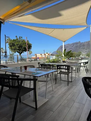 Terraza exterior de una cafetería con mesas, sillas y toldos con vistas al mar y a las montañas.