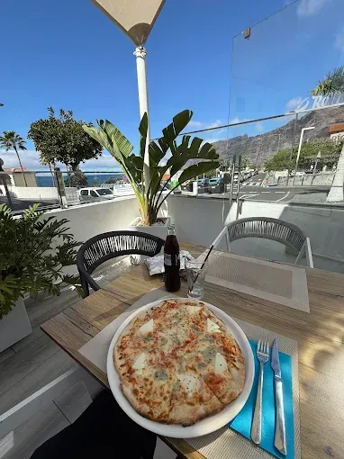 Pizza en una mesa de café junto al mar, con una bebida y cubiertos azules bajo un toldo soleado.