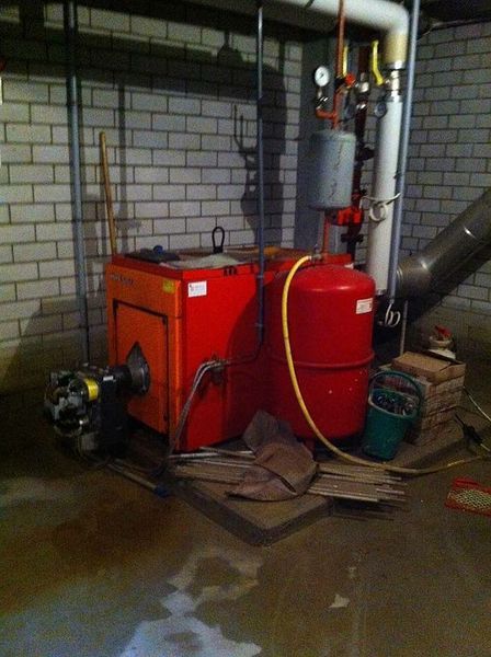 Roter und orangefarbener Ofen in einem Keller mit Rohren und Messgeräten vor einer Ziegelwand.