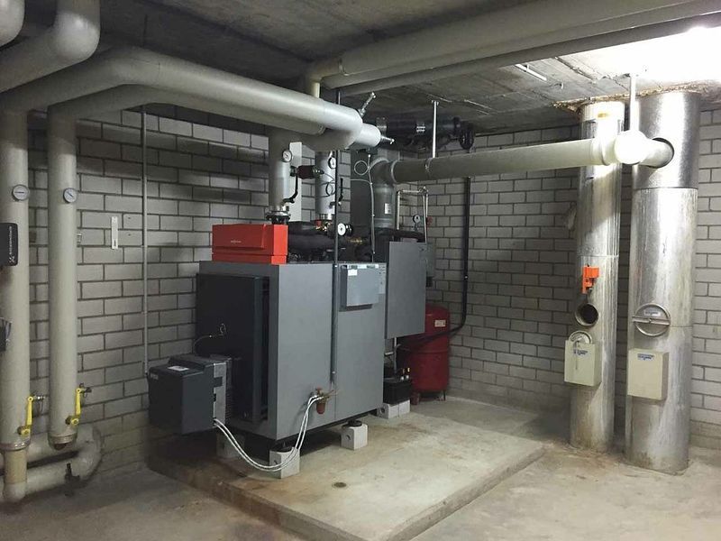 Heizungssystem in einem Keller, einschließlich Rohren, einem Kessel und einem roten Tank. Grau- und Weißtöne.