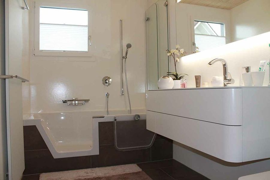 Modernes Badezimmer mit begehbarer Badewanne und schwebendem Waschtisch. Weiß- und Brauntöne; mit Fenstern und Pflanzen.