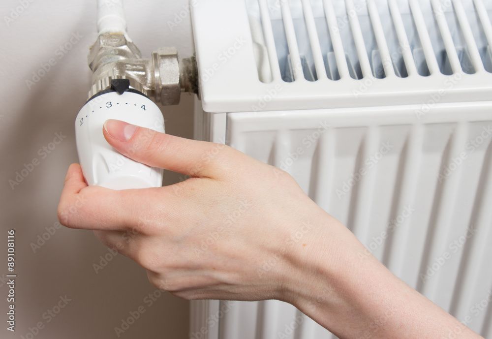 Hand dreht einen weißen Thermostatknopf an einem weißen Heizkörper im Innenbereich.