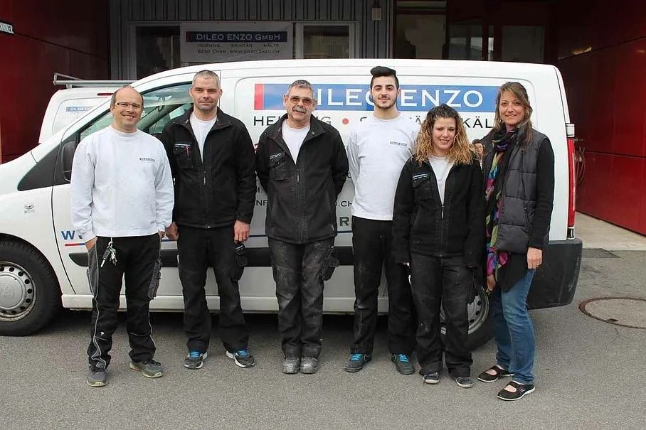 Eine Gruppe von sieben Personen steht vor einem Lieferwagen. Sie sind unterschiedlich gekleidet und stehen vor einem Gebäude.