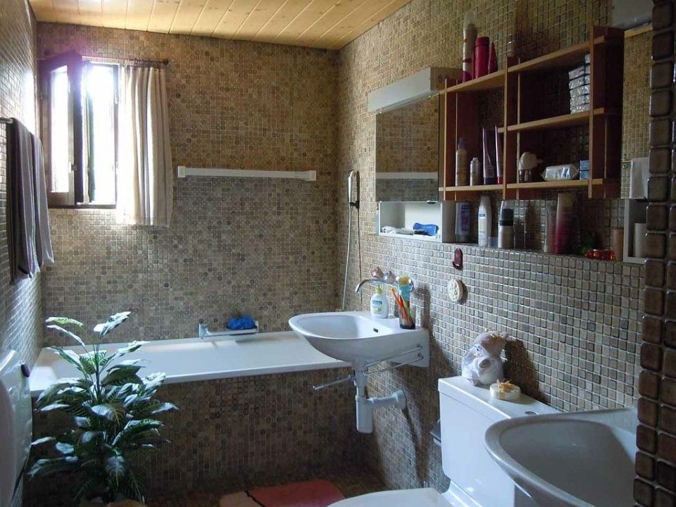 Badezimmer mit gefliesten Wänden, Badewanne, Waschbecken, Toilette und Holzregalen mit Toilettenartikeln.