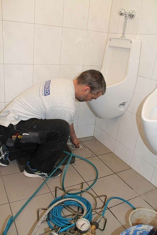 Mann hockt in einer öffentlichen Toilette und inspiziert den Sockel eines Urinals. Auf dem Boden liegen ein blauer Schlauch und Werkzeuge.