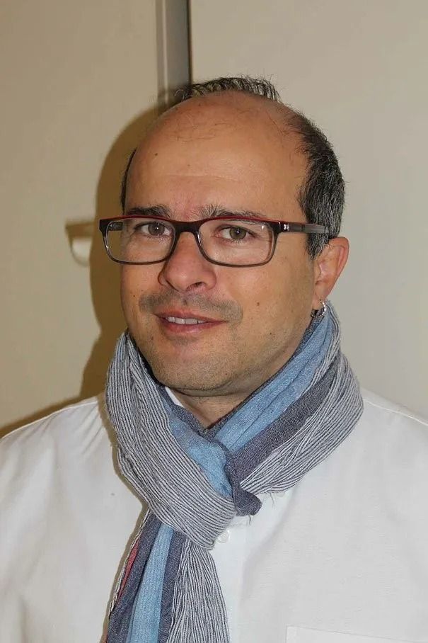 Mann mit Brille und Schal, lächelnd in einer Umgebung mit weißen Wänden.