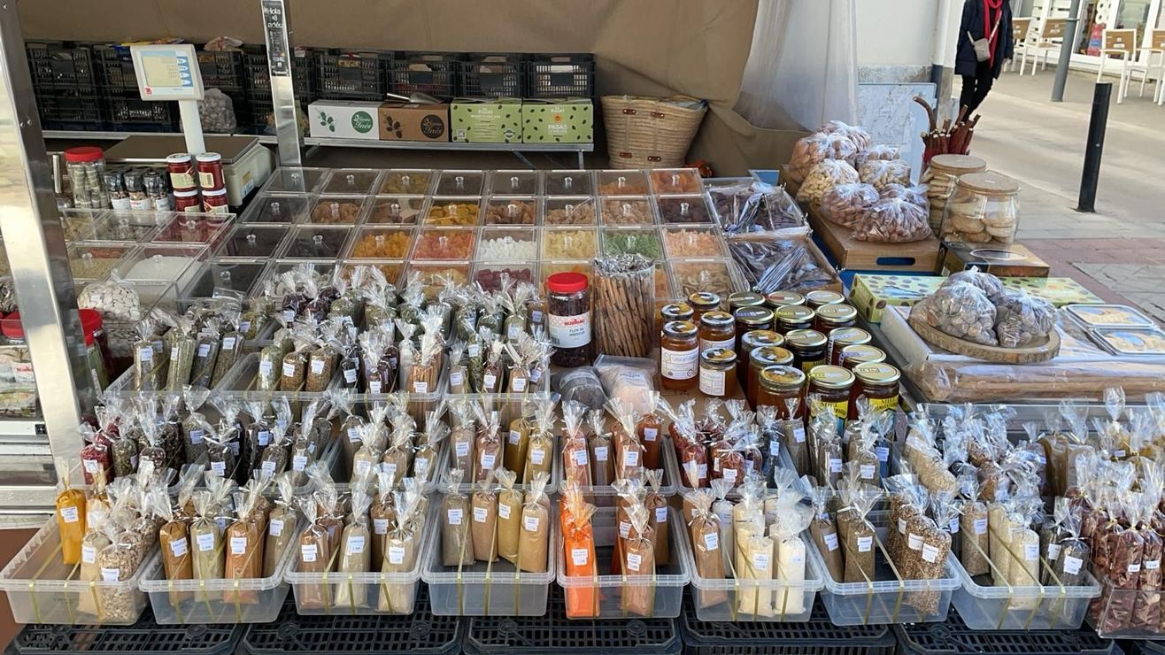 Un puesto de mercado con diversas especias, frutos secos, mermeladas y productos horneados envasados. Muchos productos se venden en envases de plástico transparente, al aire libre.