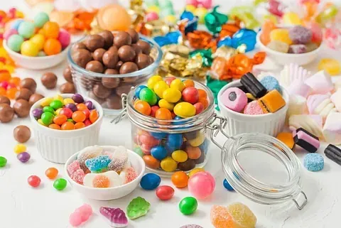 Diversos dulces de colores en tazones y frascos sobre una superficie blanca. El surtido incluye chocolates, gominolas y caramelos duros.