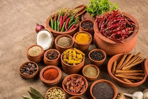 Especias variadas en pequeñas macetas de terracota, que incluyen chiles, cúrcuma, canela y granos de pimienta, dispuestos sobre una superficie marrón.