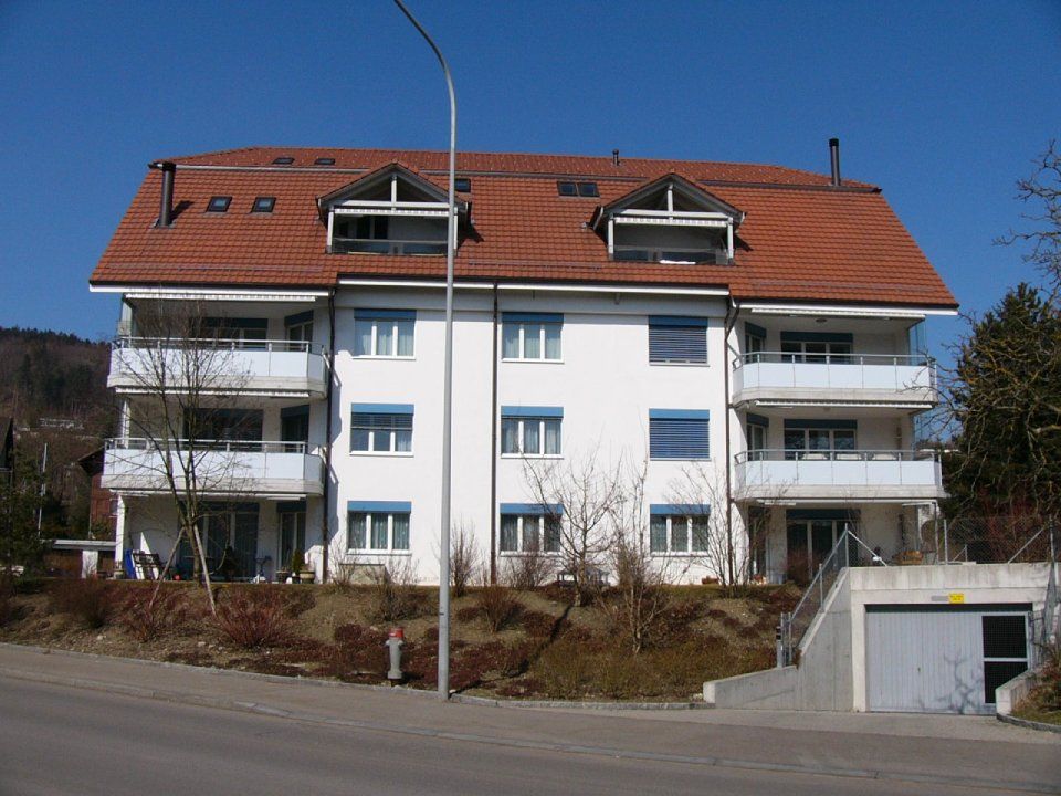 Weißes Mehrfamilienhaus mit rotem Ziegeldach, Balkonen und Garageneinfahrt. Blauer Himmel.