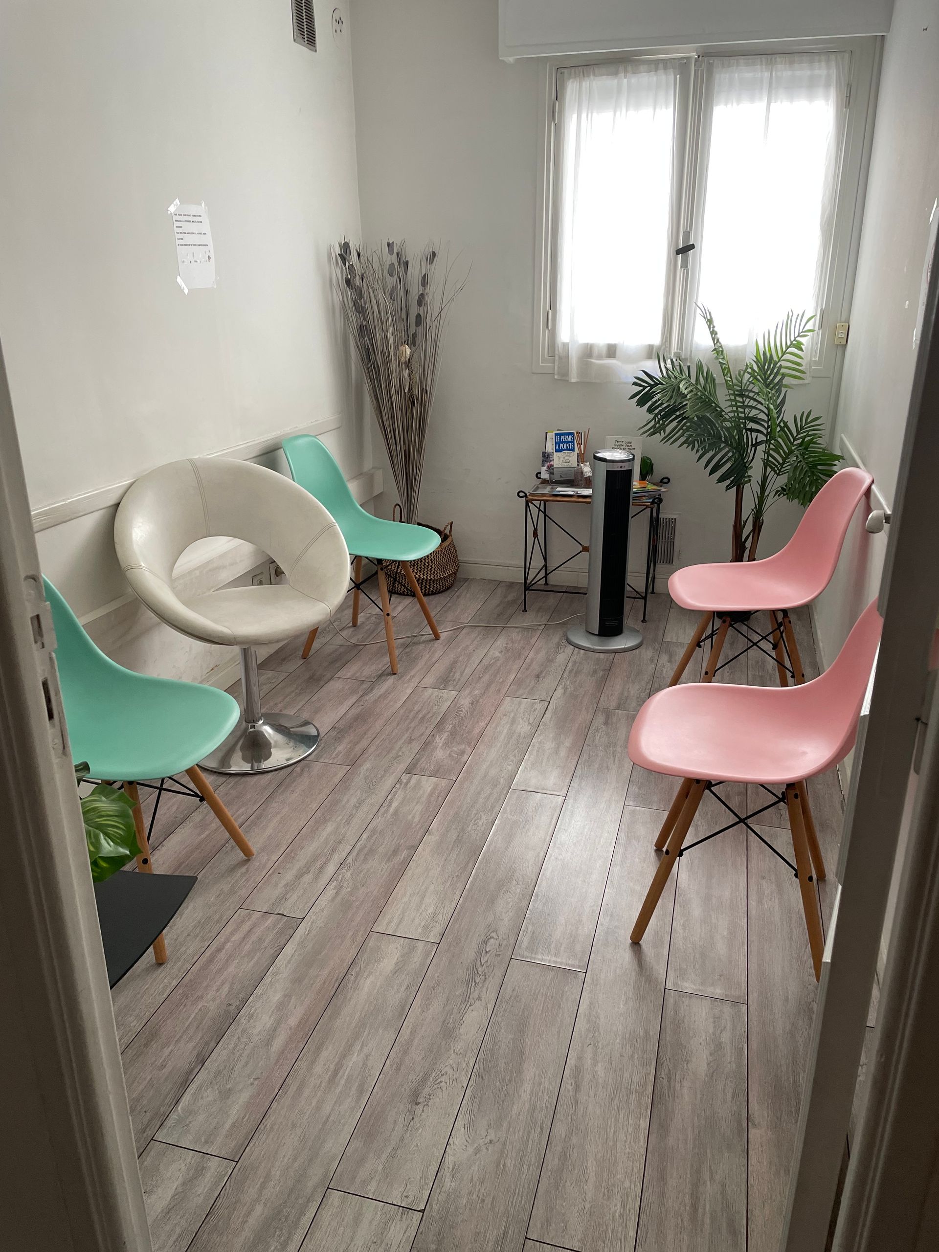 Petite salle d'attente avec des chaises pastel, un fauteuil blanc, une table d'appoint, des plantes et une fenêtre donnant sur un parquet.