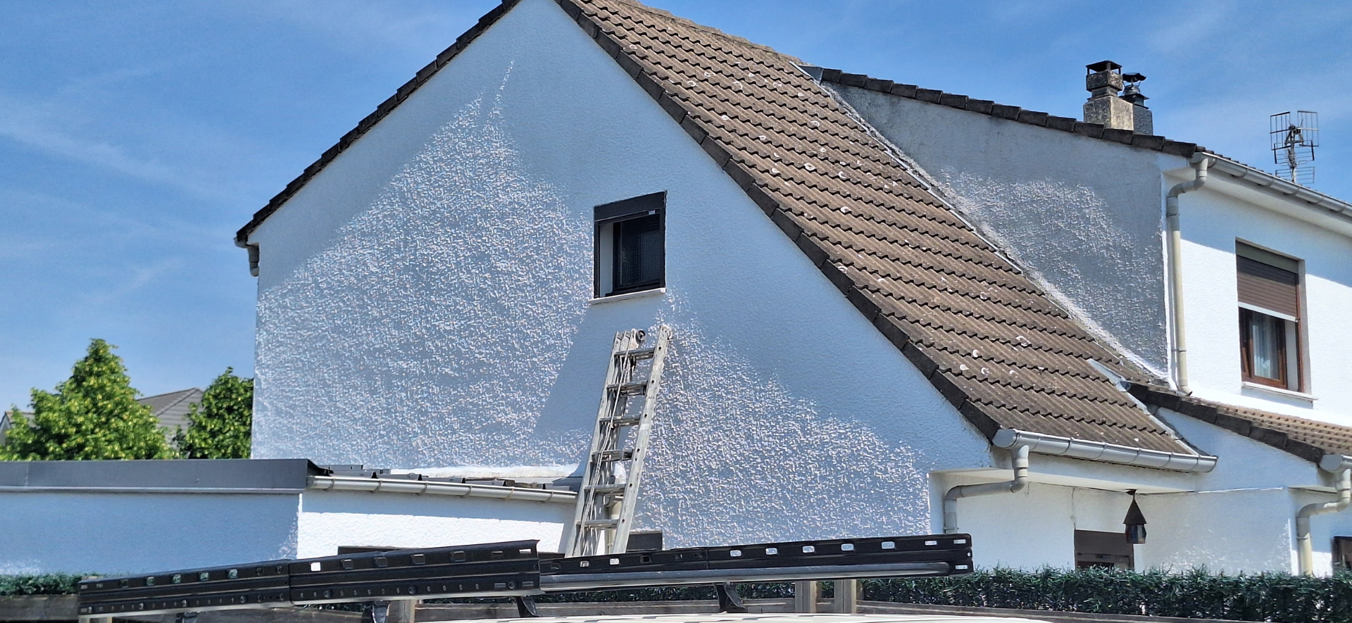 Une maison blanche au toit brun et une échelle menant à une fenêtre, sur fond de ciel bleu.