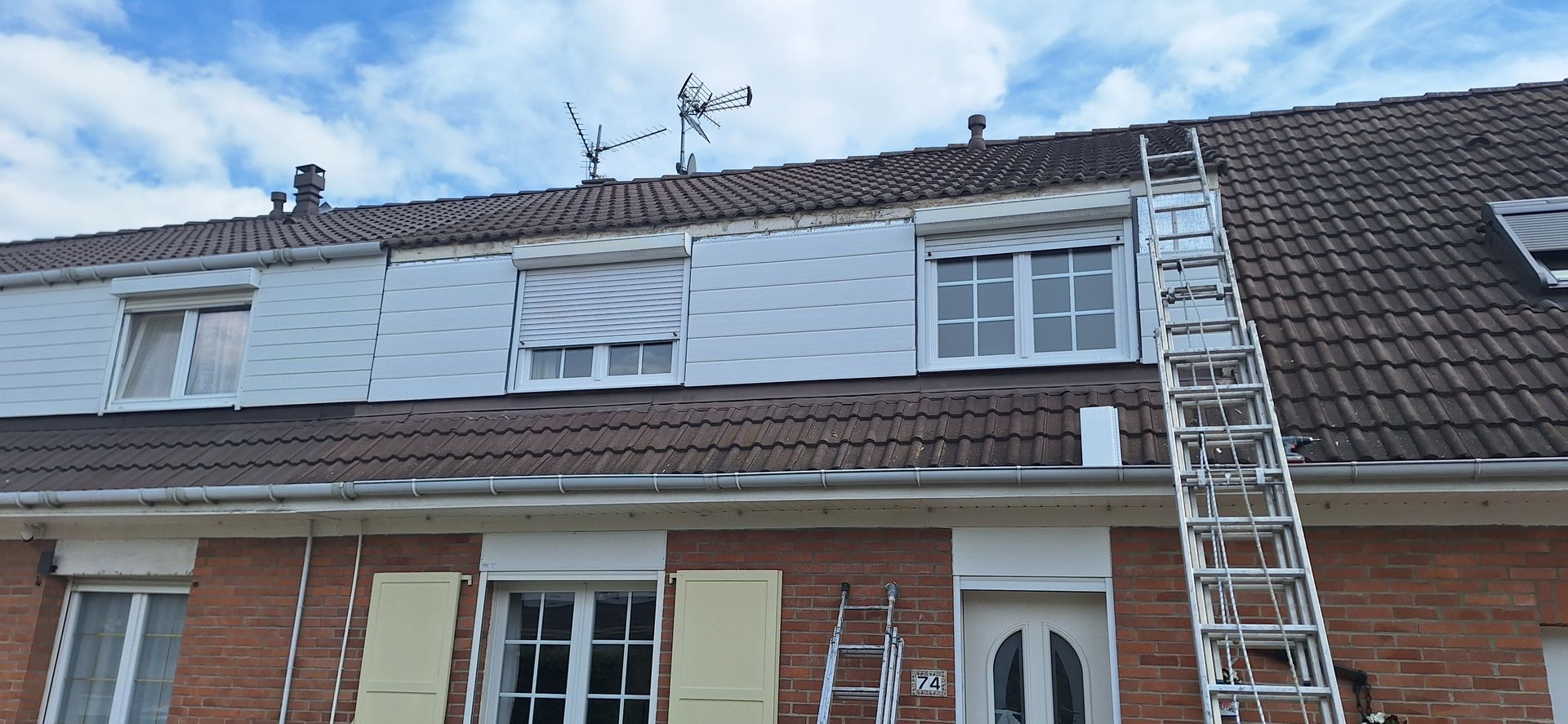 Une échelle est appuyée contre le toit d'un bâtiment en briques, dont on aperçoit les fenêtres et l'antenne.