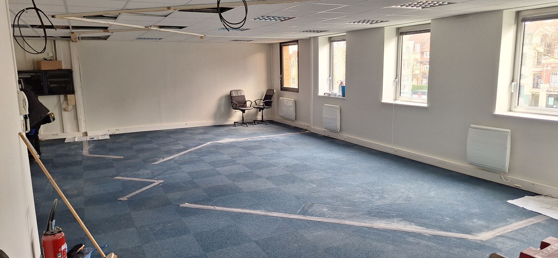 Bureau vide avec moquette bleue, murs blancs et trois fenêtres. Deux chaises sont placées près d'une fenêtre.