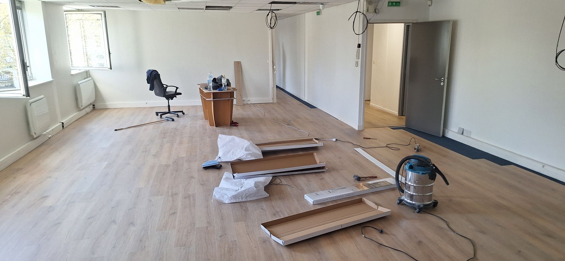 Une pièce en rénovation. Des morceaux de bois et un aspirateur jonchent le sol. Un bureau et une chaise se trouvent au loin.