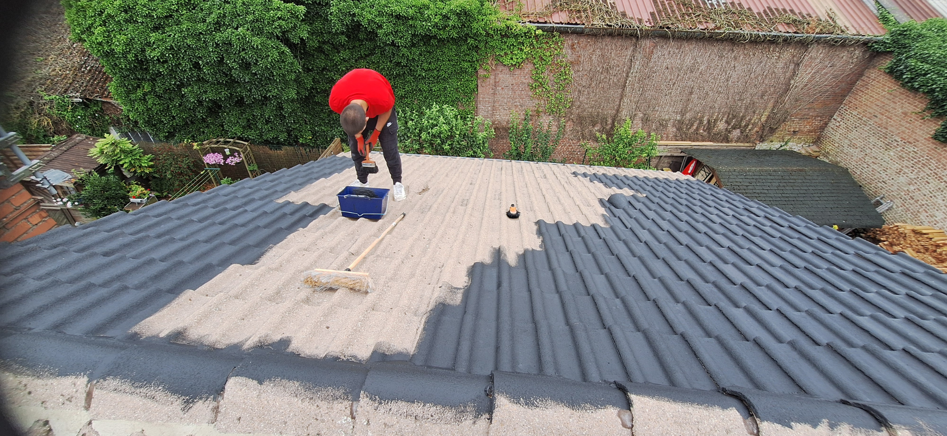 Une personne en chemise rouge travaille sur un toit gris avec des outils, entourée de feuillage vert et d'un mur de briques.