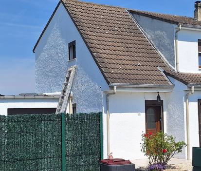 Maison blanche au toit marron, échelle appuyée contre le mur. Clôture verte et ciel bleu en arrière-plan.
