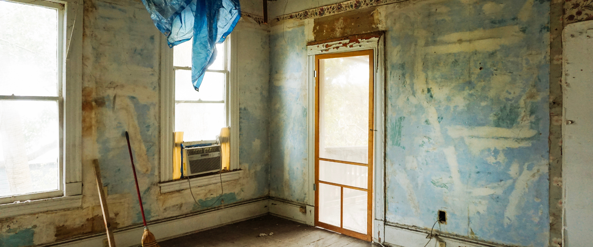 Intérieur d'une pièce avec de la peinture bleue écaillée, une porte moustiquaire ouverte, des fenêtres et des murs apparents.