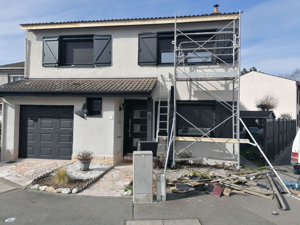 Maison à deux étages avec volets noirs, garage et échafaudages pour rénovation. Façade grise, débris devant.