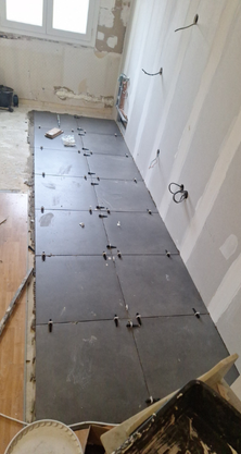 Pose de carrelage en cours : carreaux gris foncé posés avec des croisillons dans une pièce étroite, près d’une fenêtre