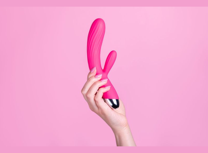 Une main tient un vibromasseur en silicone rose sur un fond rose uni.