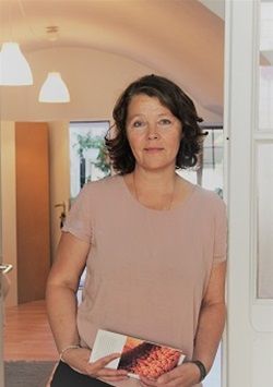 Frau mit kurzen braunen Haaren, rosa Hemd und einem Buch in der Hand. Sie steht mit neutralem Gesichtsausdruck in einem Hauseingang.