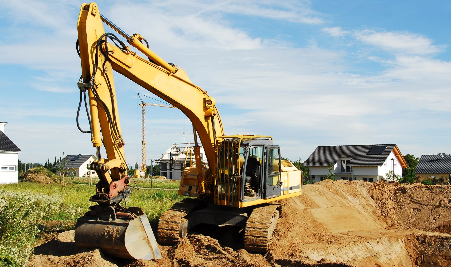 Excavatrice jaune creusant dans un tas de terre, maisons et ciel bleu en arrière-plan.