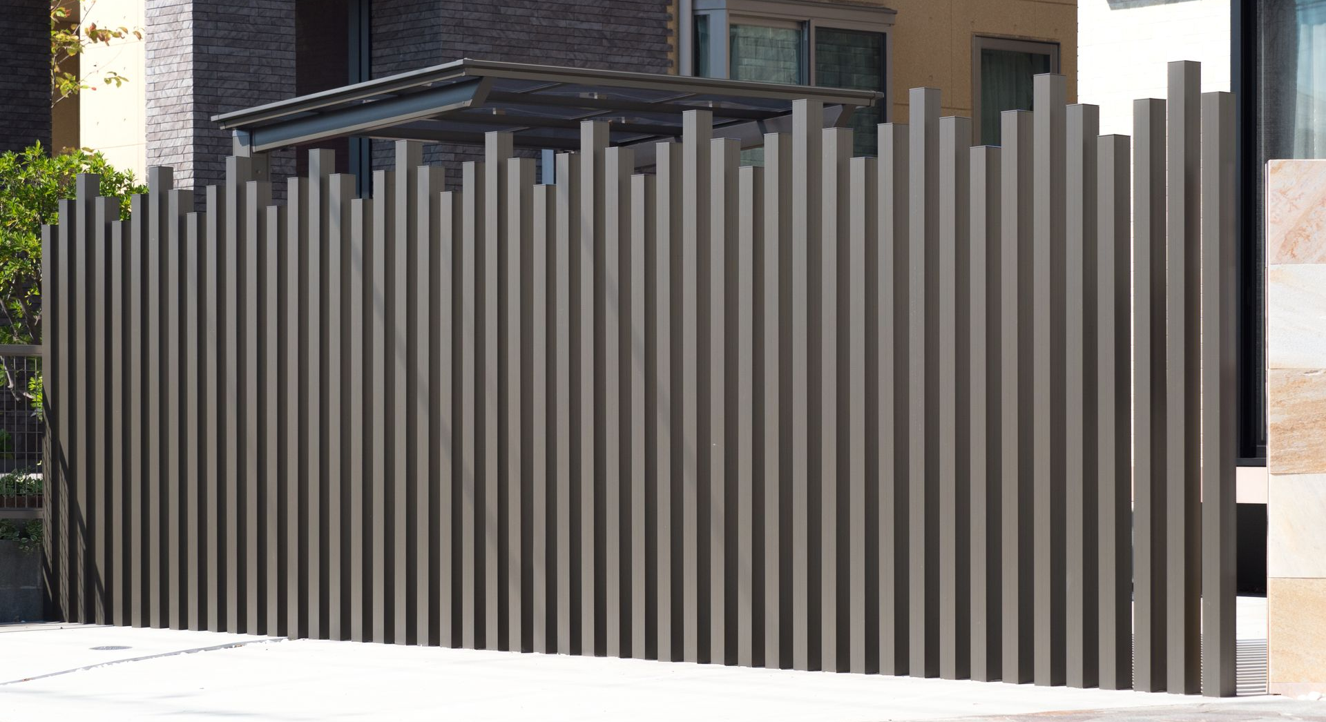 Clôture à lattes verticales grises devant un bâtiment, projetant des ombres.