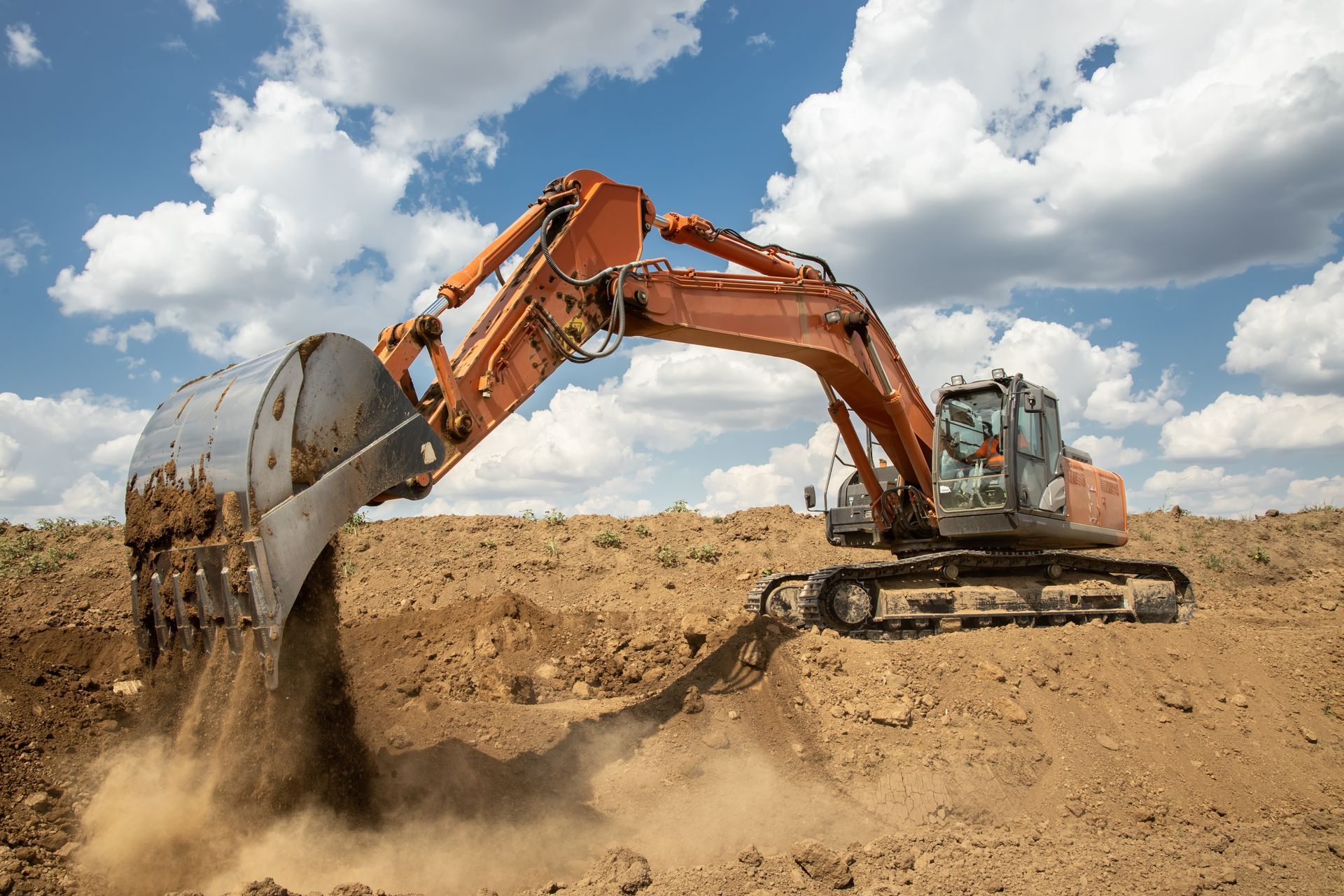Excavatrice orange creusant dans un sol brun sous un ciel bleu nuageux.