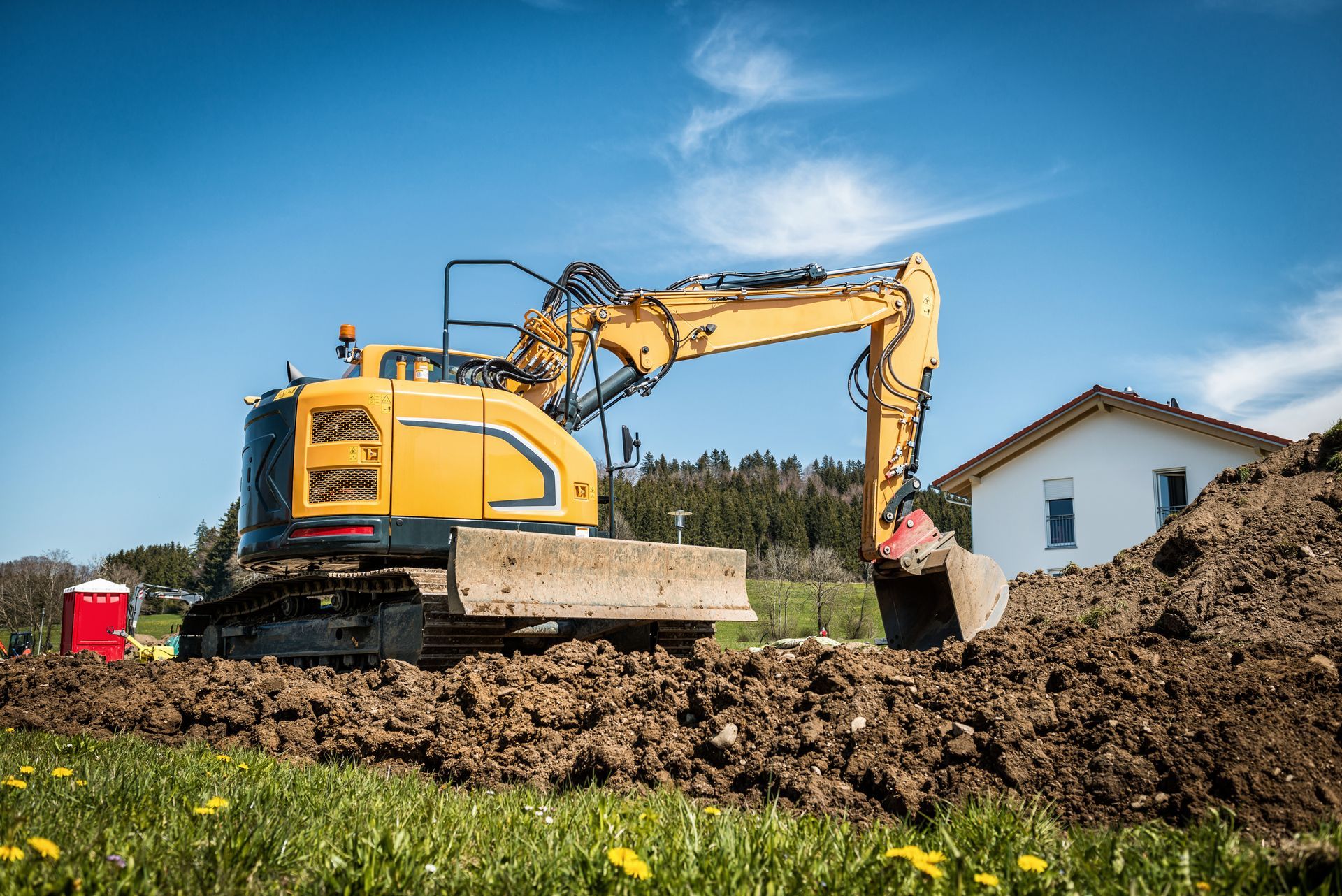 Excavatrice jaune creusant une tranchée sur un chantier herbeux près d'une petite maison blanche sous un ciel bleu.