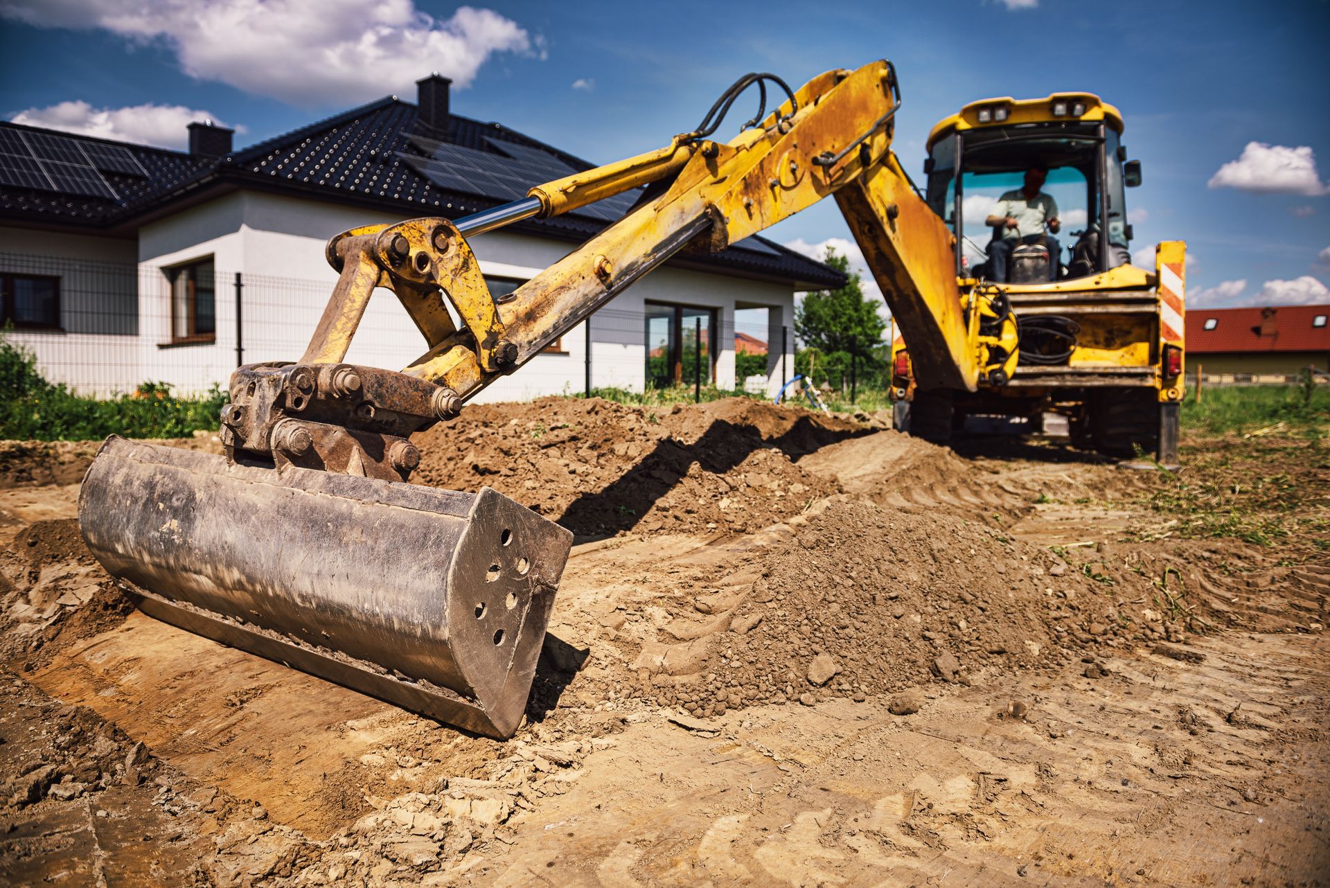 Excavatrice jaune creusant dans une zone de terre devant une maison par une journée ensoleillée.