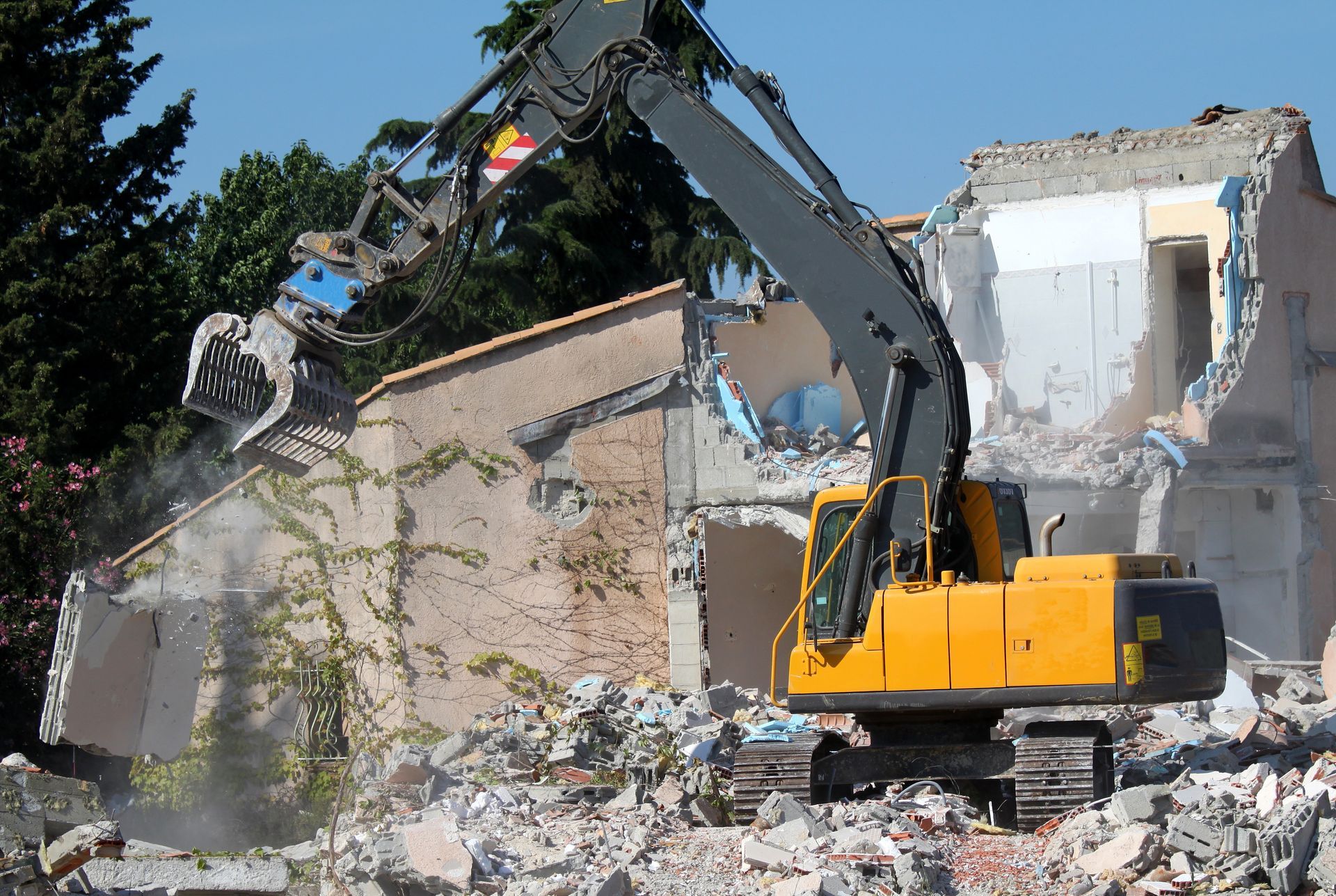 Excavateur jaune démolissant un bâtiment ; décombres, ciel bleu, arbres en arrière-plan.