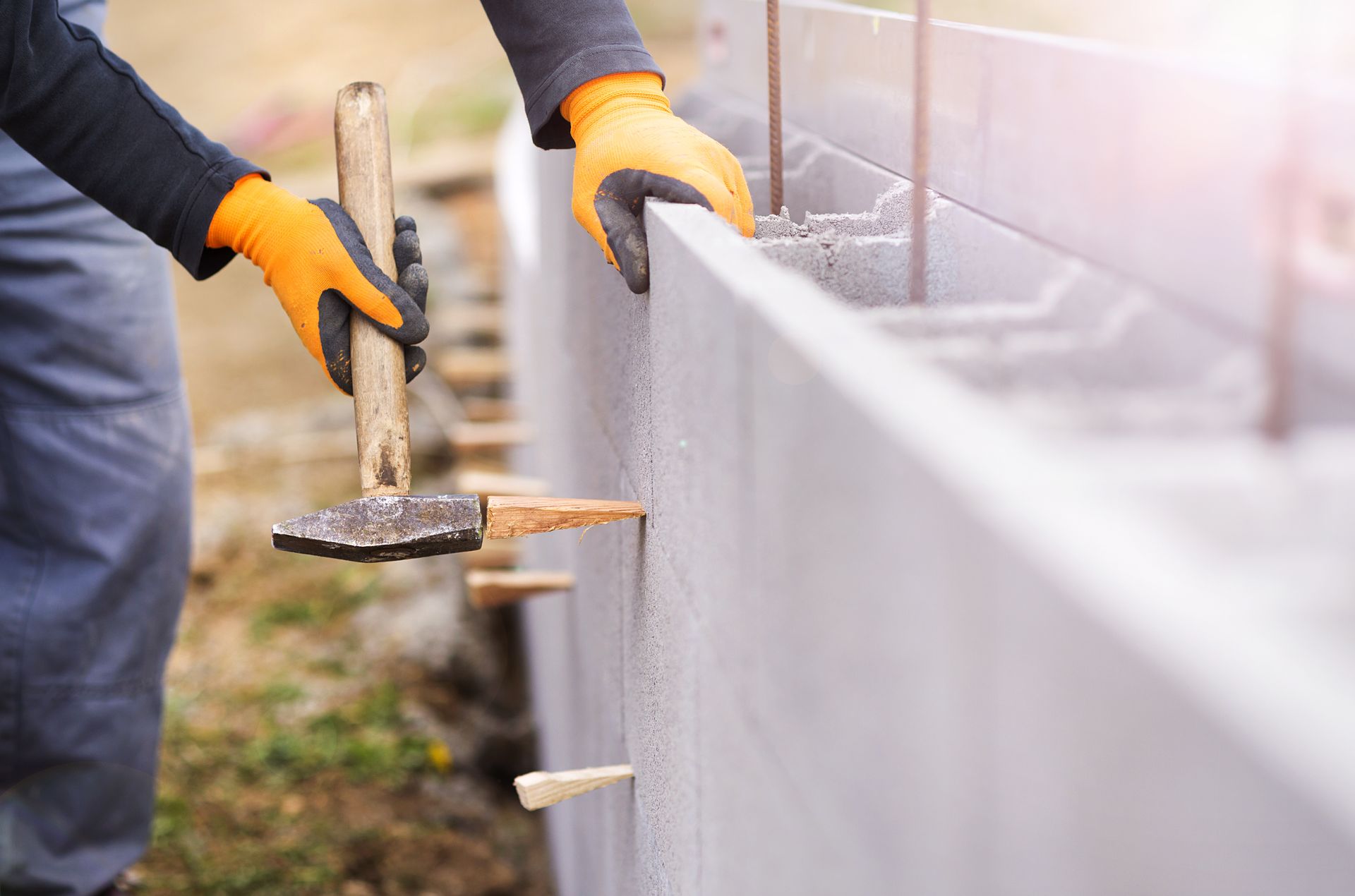 Une personne portant des gants orange utilise un marteau pour construire un mur de parpaings à l'extérieur.