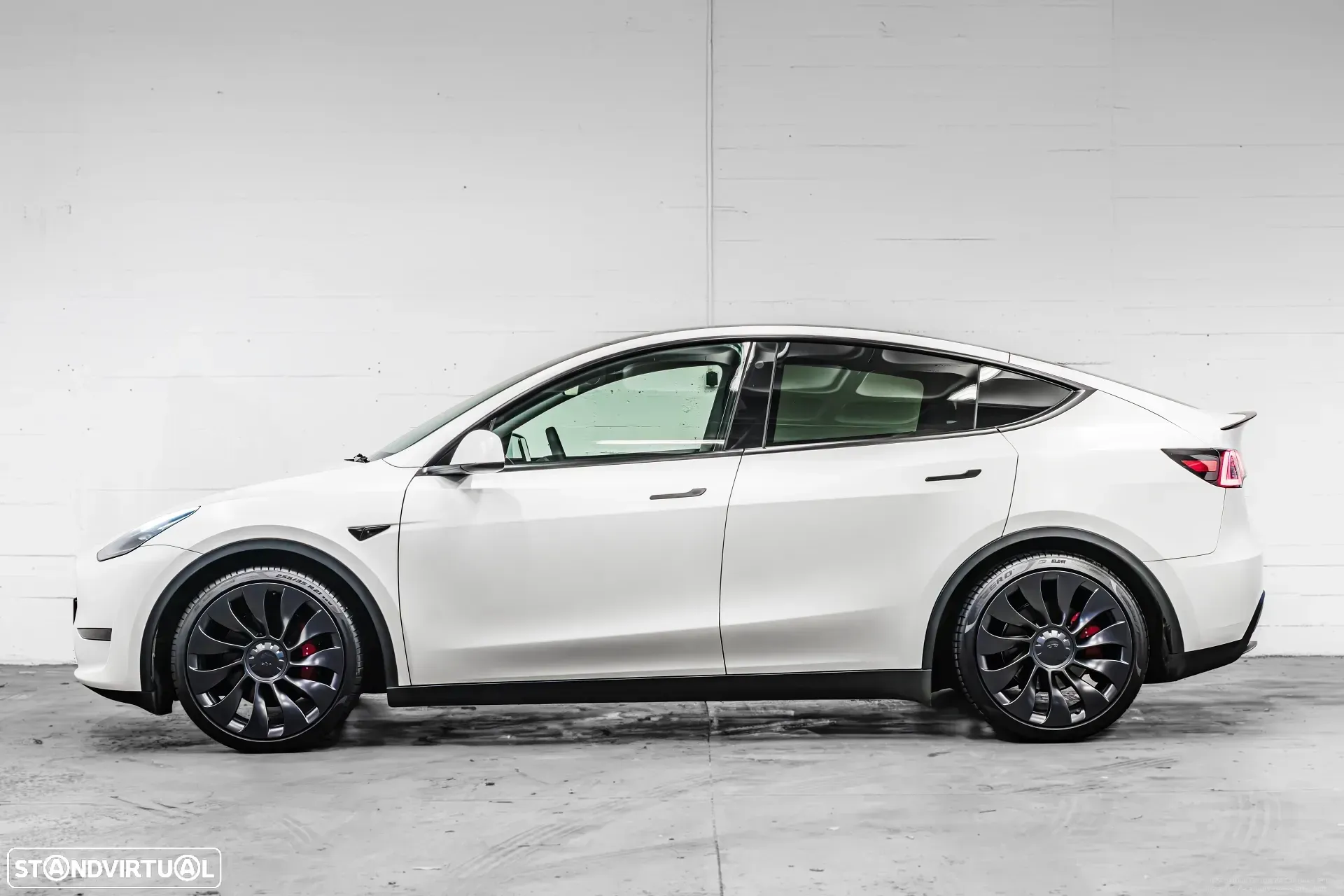 SUV Tesla Model Y branco com rodas pretas estacionado contra uma parede branca.