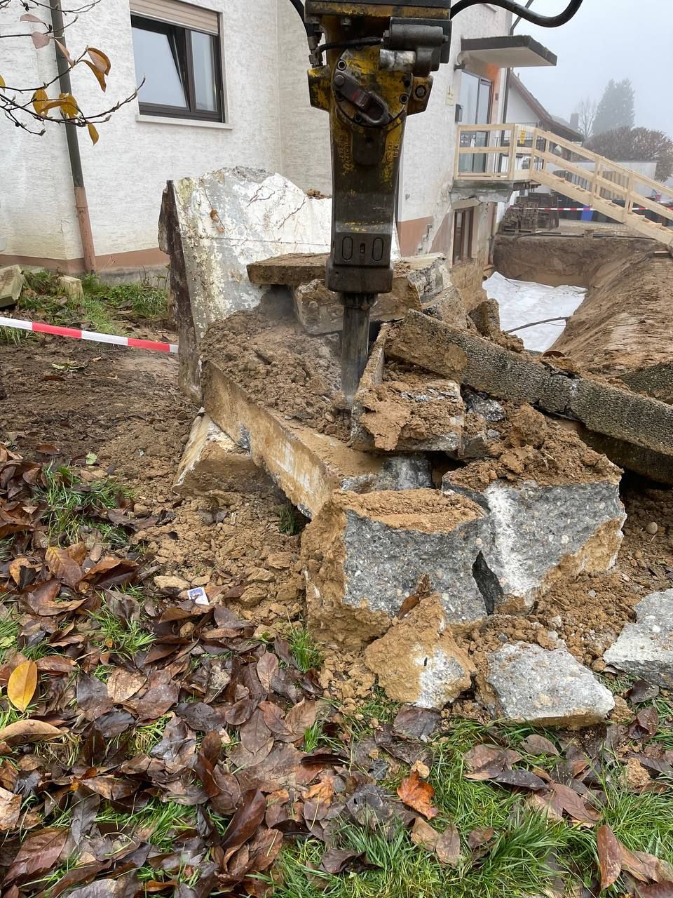 Ein Baggerhammer zerkleinert einen Steinblock neben einem Gebäude; Schuttteile liegen verstreut auf dem Boden.